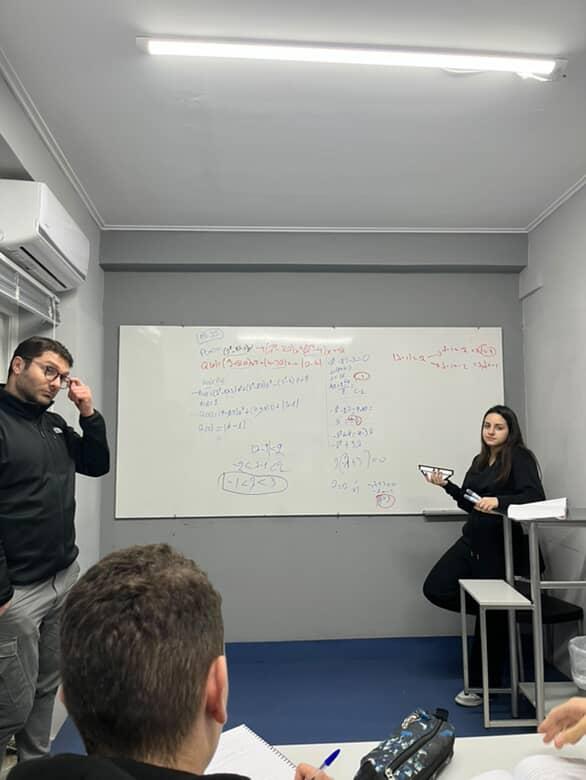 Ο Πάρης Βαρύτης σε αίθουσα διδασκαλίας, ενώ μια μαθήτρια λύνει άσκηση στον πίνακα και άλλοι μαθητές κρατούν σημειώσεις.